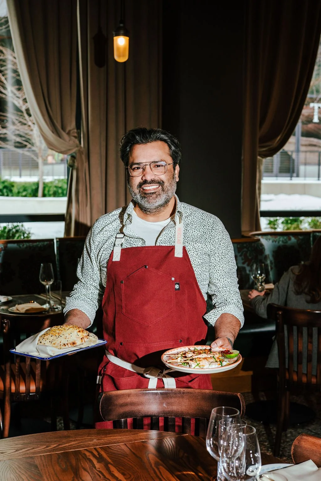 A man wearing glasses and a red apron stands in a restaurant holding a plate of food in each hand. The restaurant has dark curtains, large windows, and wooden furniture.