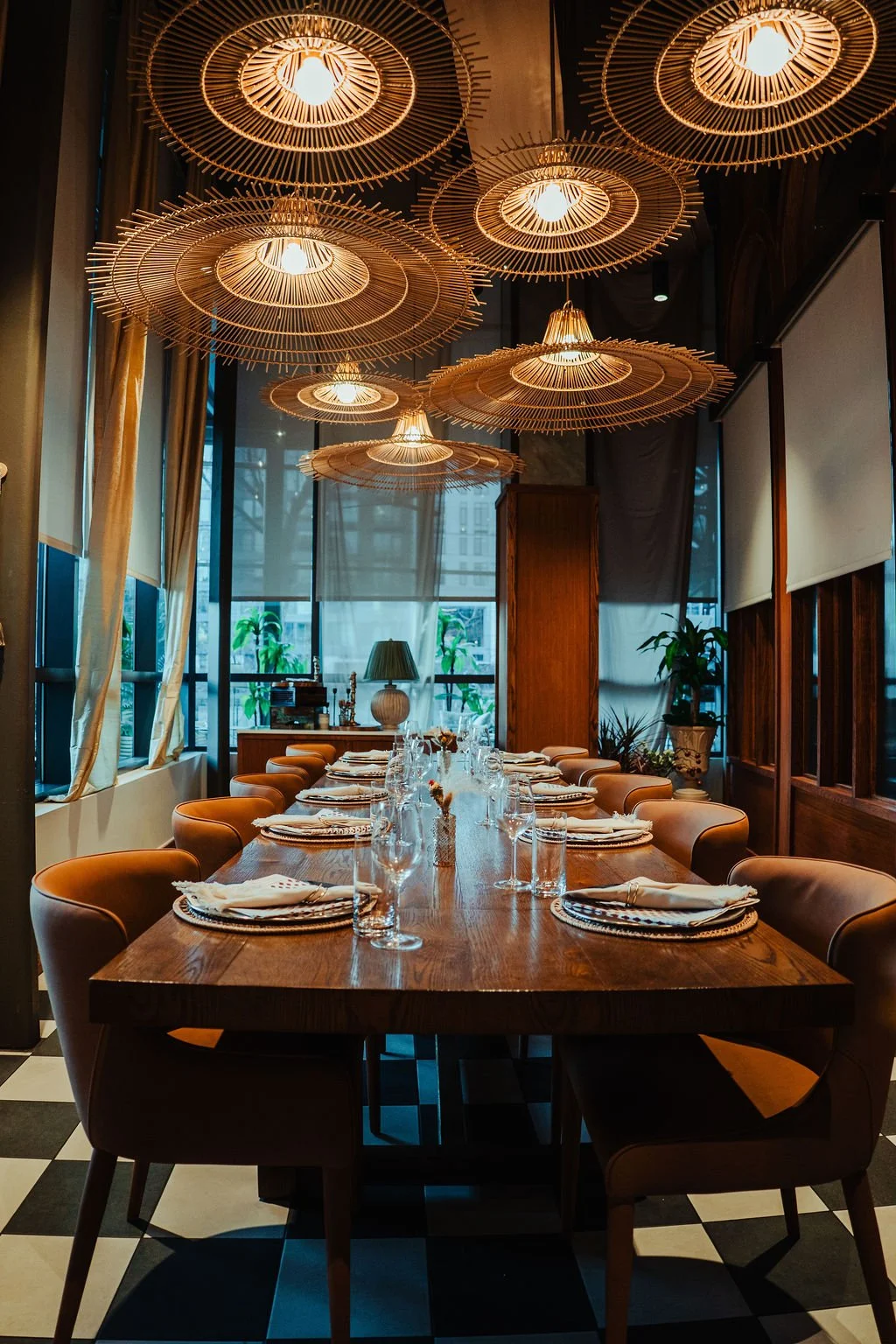 A decorated dining room with a large wooden table set for a meal, surrounded by chairs. The ceiling features multiple large, circular, wooden pendant lights. The room has large windows with drapes, and there are several potted plants inside.