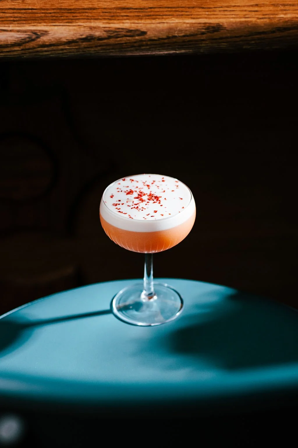 A cocktail glass filled with a pink drink, topped with white foam and red sprinkles, placed on a blue surface.
