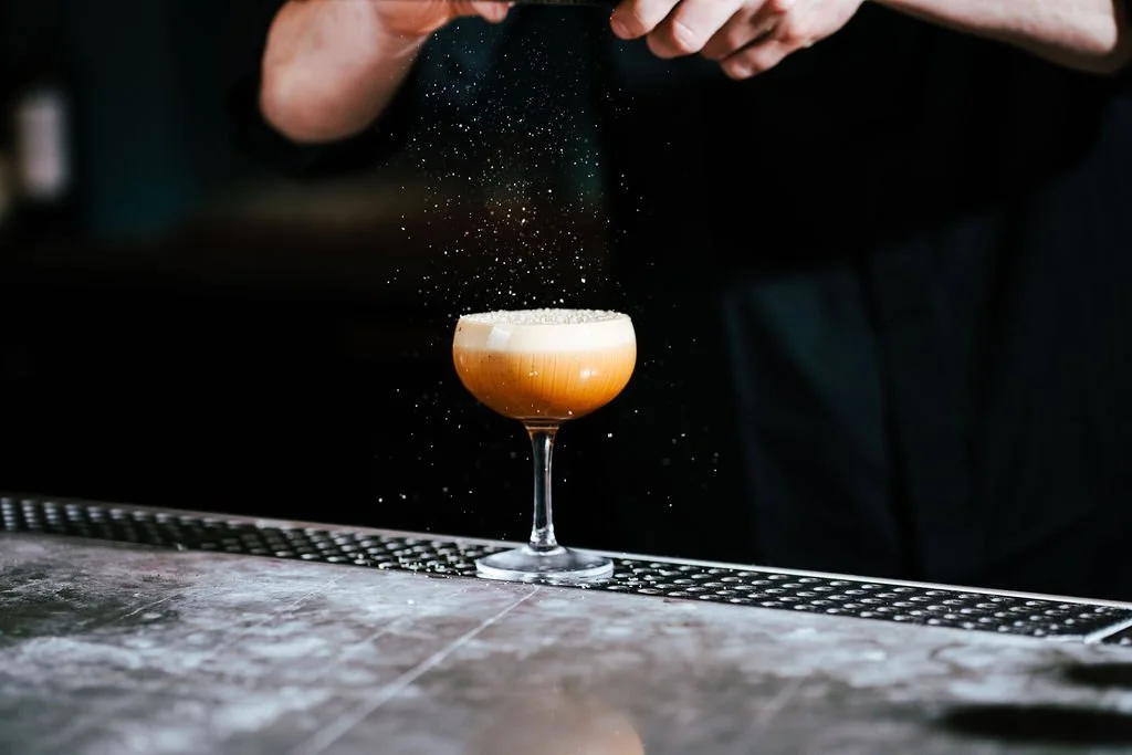 A bartender dusting powdered sugar over a cocktail glass filled with a layered coffee-colored drink, set on a bar counter.
