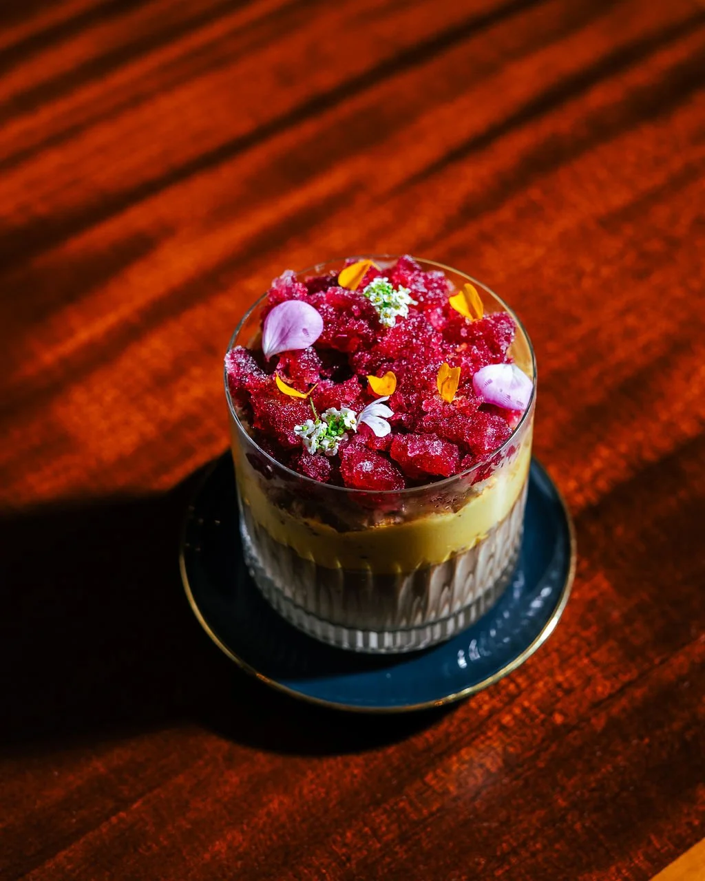 A dessert topped with crushed berries, edible flower petals, and small white flowers in a glass on a dark plate, set on a wooden table.