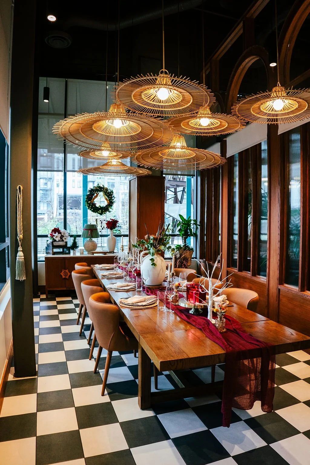 A festive dining room decorated for Christmas with a long wooden table set with napkins, vases, and holiday ornaments, illuminated by a modern wooden chandelier, and featuring a Christmas wreath and poinsettias in the background.