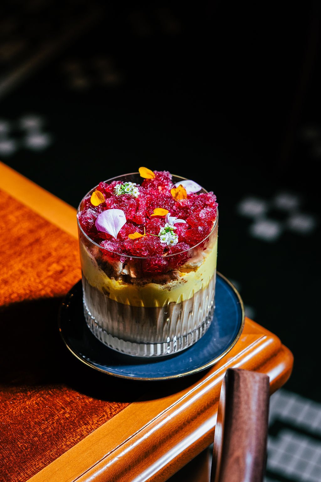 A layered dessert in a glass cup topped with edible flowers and garnishes, sitting on a blue saucer on a wooden table.