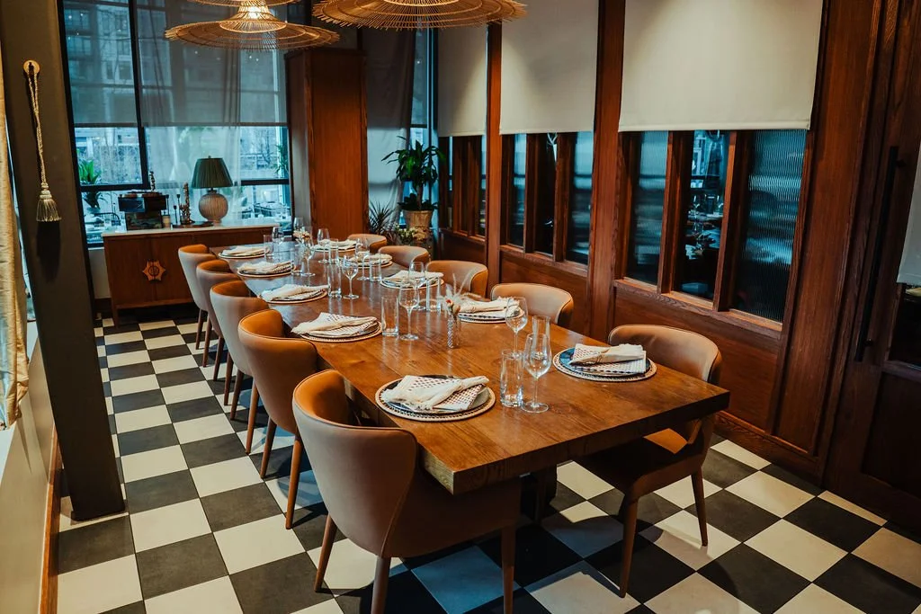 A long wooden dining table set for a meal, with beige chairs, glassware, and napkins, in a cozy restaurant or private dining room with checkered black and white floor, large windows, and warm lighting.