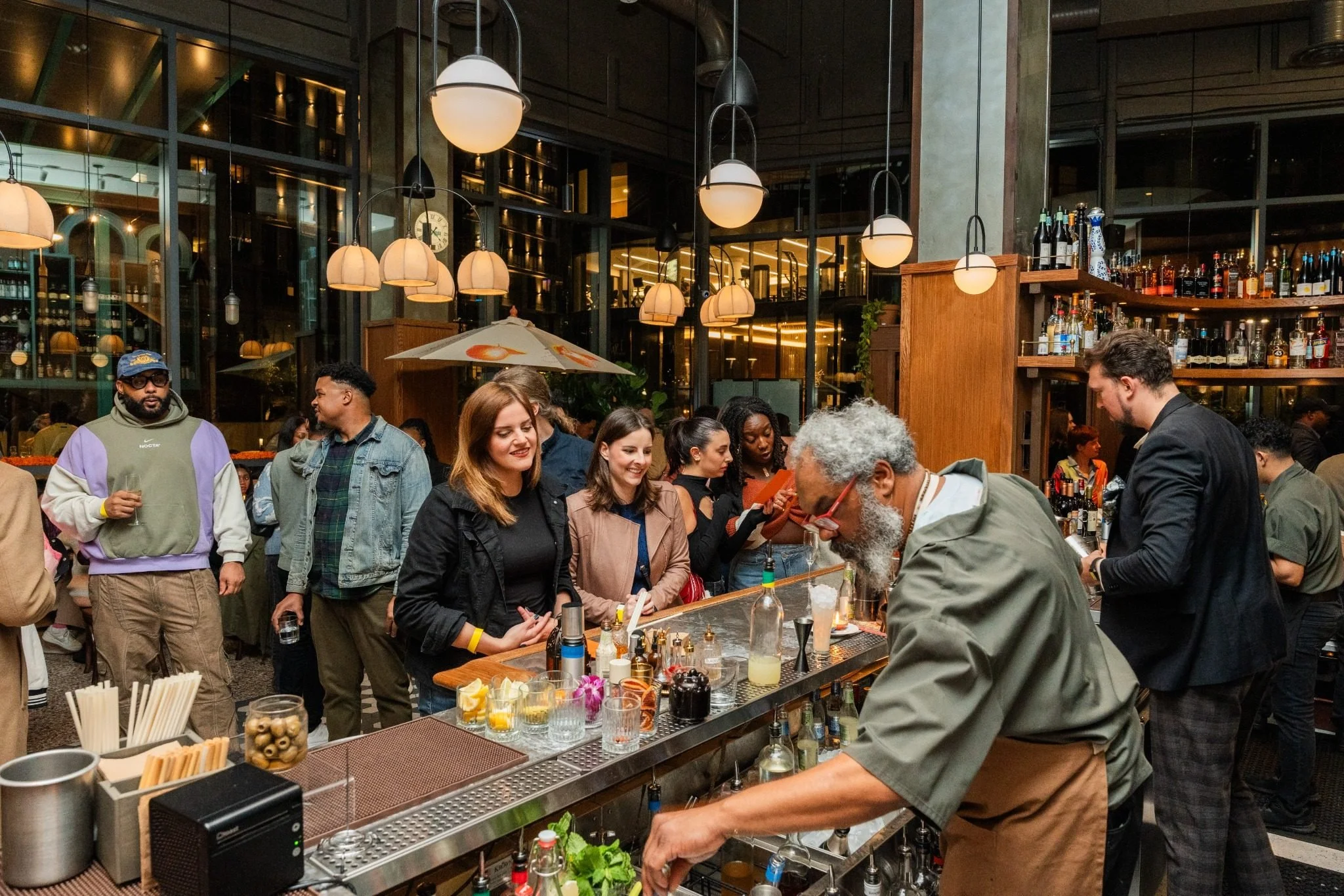 People at a bar ordering drinks at Karravaan private event. Bartenders preparing crafted cocktails in a modern venue with warm lighting and shelves of bottles.