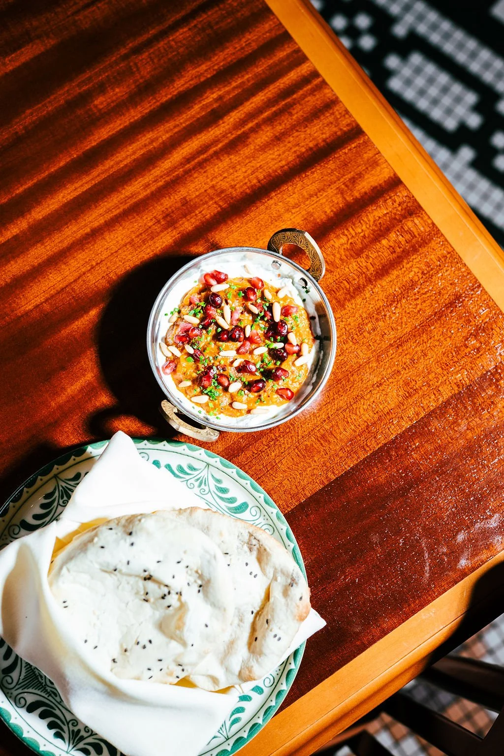 A bowl of Indian curry garnished with pomegranate seeds and pine nuts, served alongside a piece of naan bread in a decorative bowl on a wooden table.