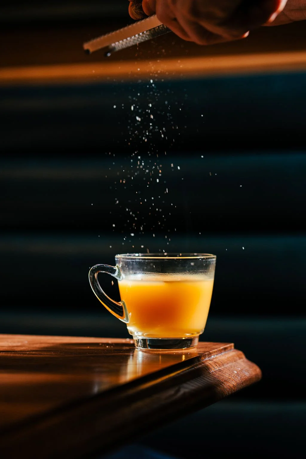 A cup of coffee on a wooden table with someone sprinkling sugar into it.