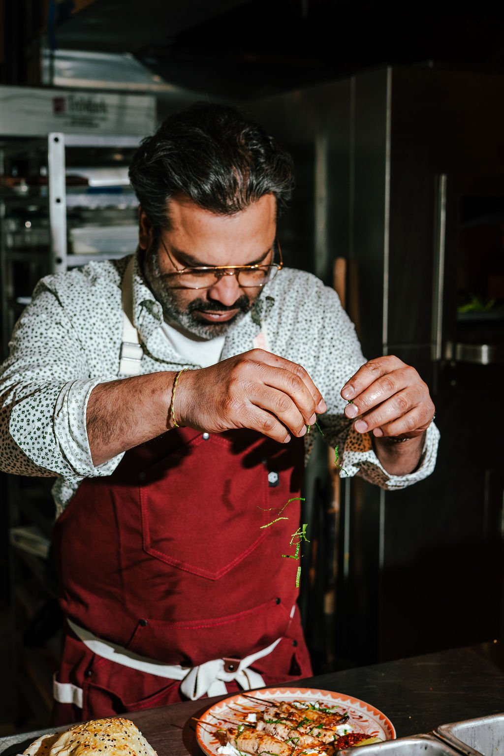 A man in a red apron and glasses is garnishing a dish in a professional kitchen.