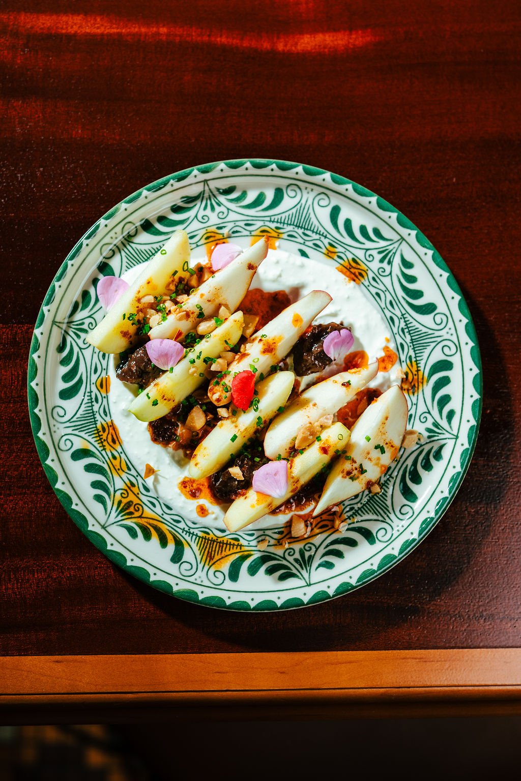 A plate of Mexican food with sliced white vegetables, topped with chopped nuts, herbs, flower petals, and sauce, on a decorative green and white patterned plate on a dark wooden table.