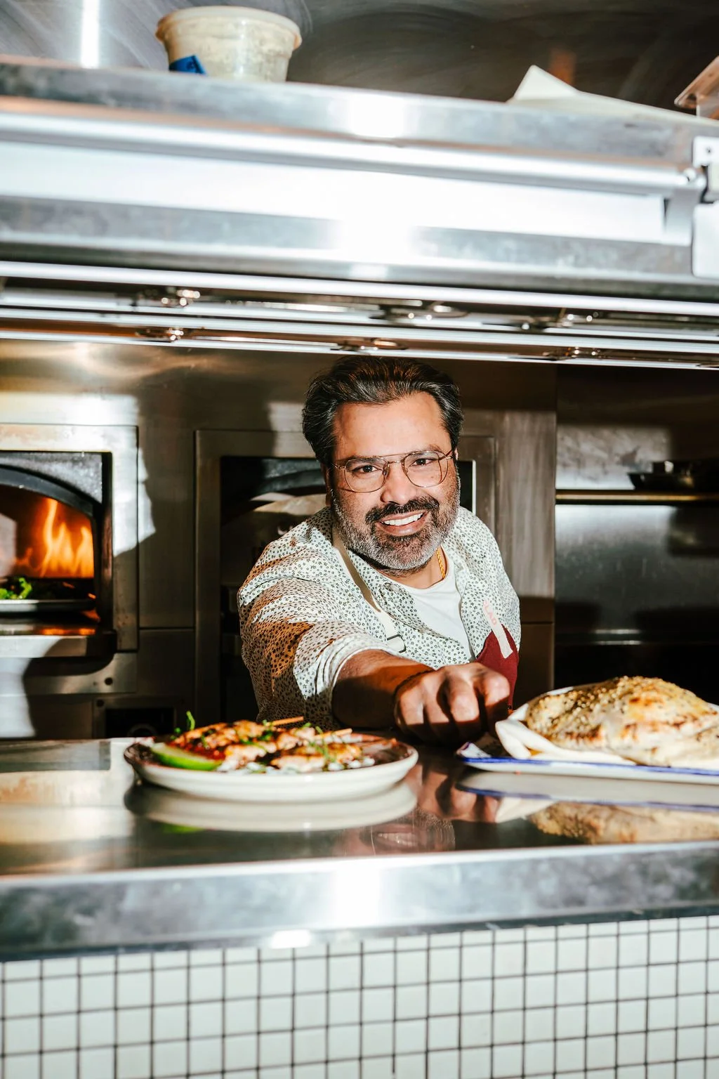 A man in a patterned shirt and glasses is smiling in a restaurant kitchen, serving food from the counter. There are plates with food, a pizza on a white tray, and an oven with a visible flame in the background.