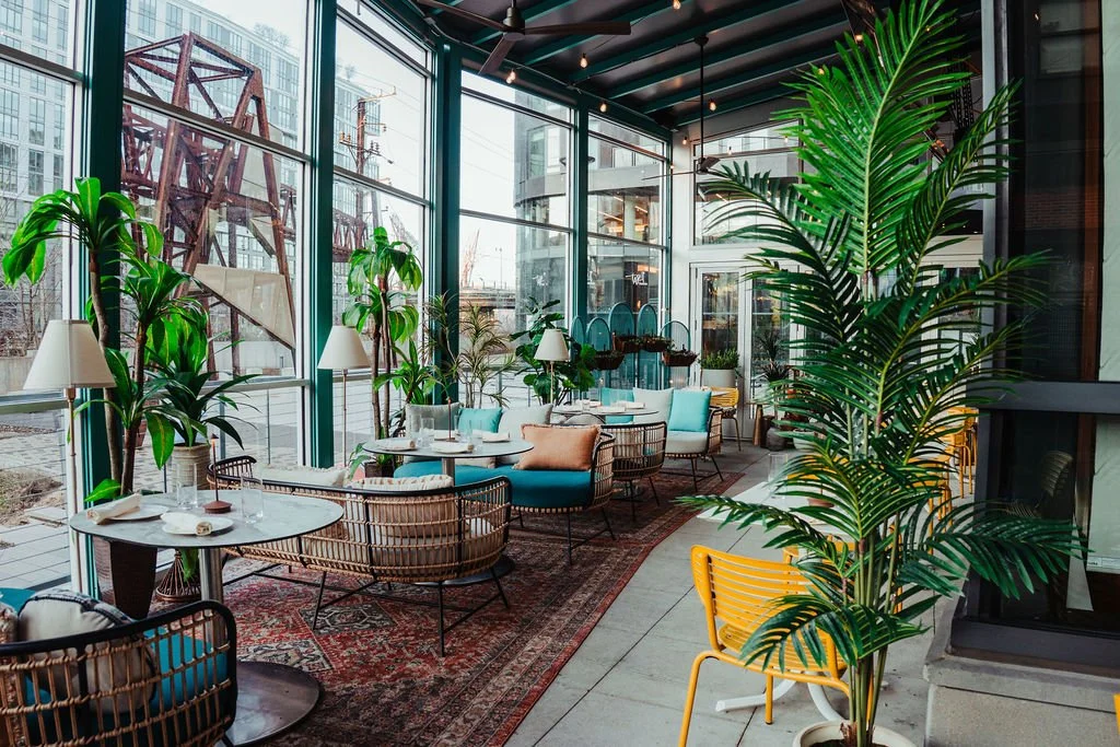 Sunlit indoor restaurant or cafe with glass walls, tropical plants, colorful chairs, and tables set for dining.
