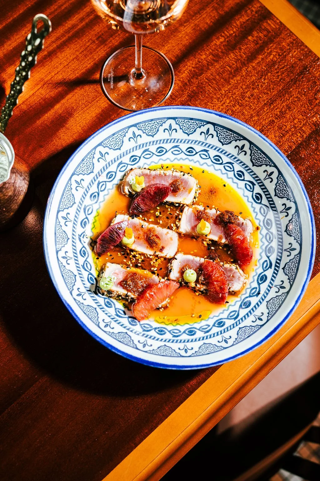 Sashimi-style tuna with blood orange slices, garnished with sesame seeds and green sauce, served in a patterned blue and white bowl.