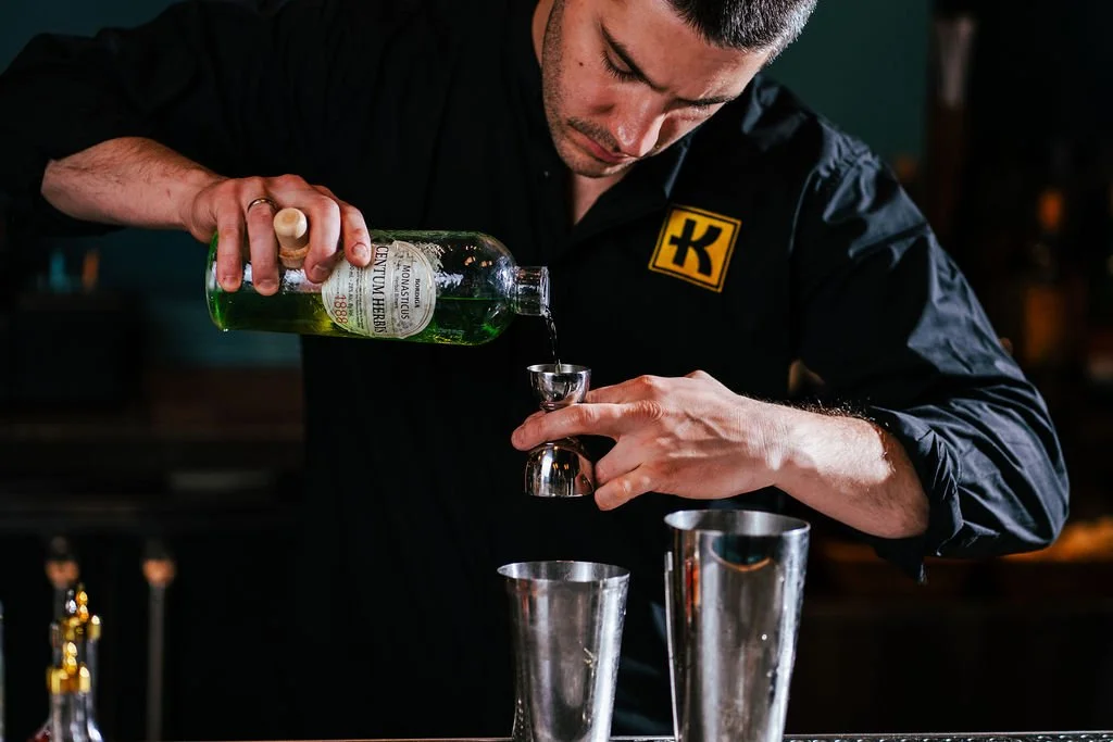A bartender pouring green liquid from a bottle into a jigger, with two metal shakers on the bar.