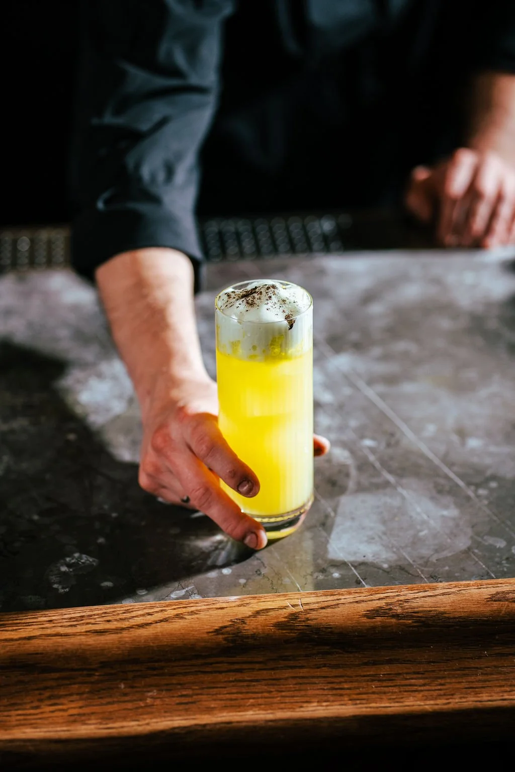 A bartender in a black shirt presenting a tall glass of yellow cocktail with whipped cream and cocoa topping on a bar counter.