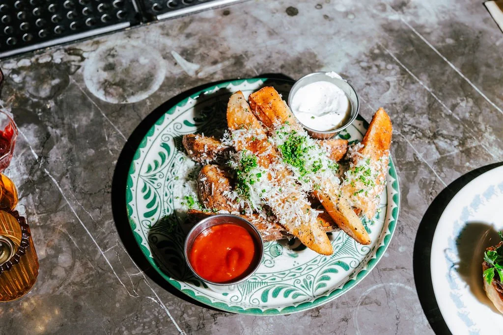 Plate of potato wedges topped with shredded cheese, garnished with chopped chives, served with side of sour cream and ketchup.