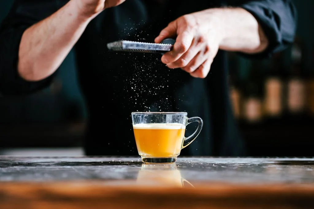A person sprinkling powdered ingredient into a glass cup of yellow liquid on a table.