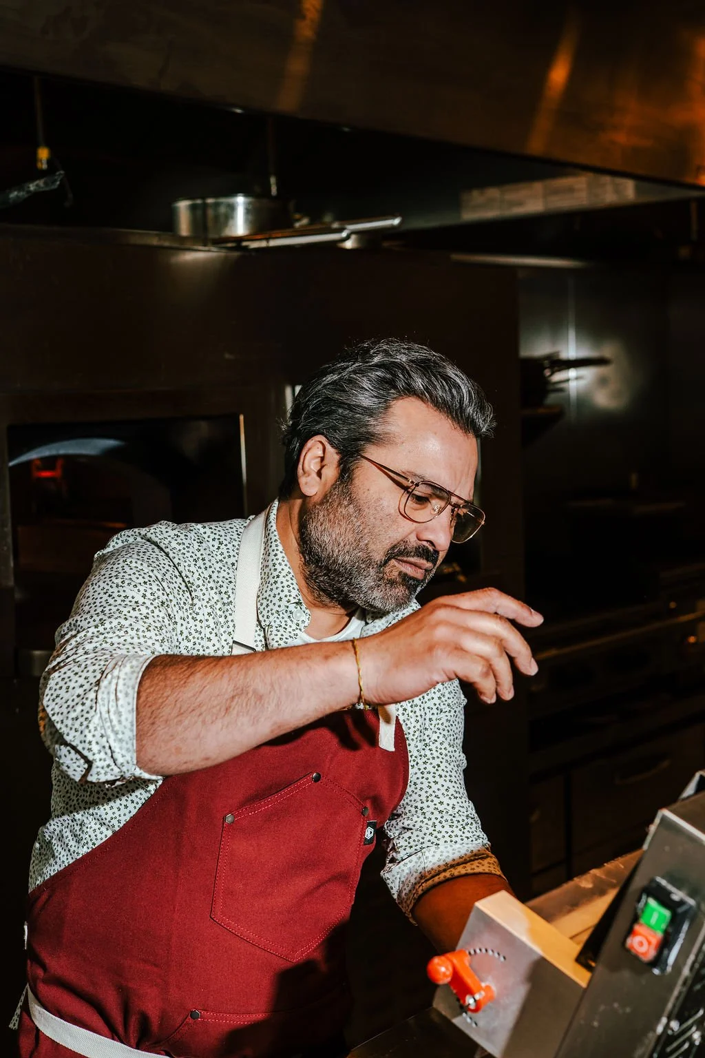 A man with glasses and a beard, wearing a white patterned shirt and a red apron, working in a professional kitchen.