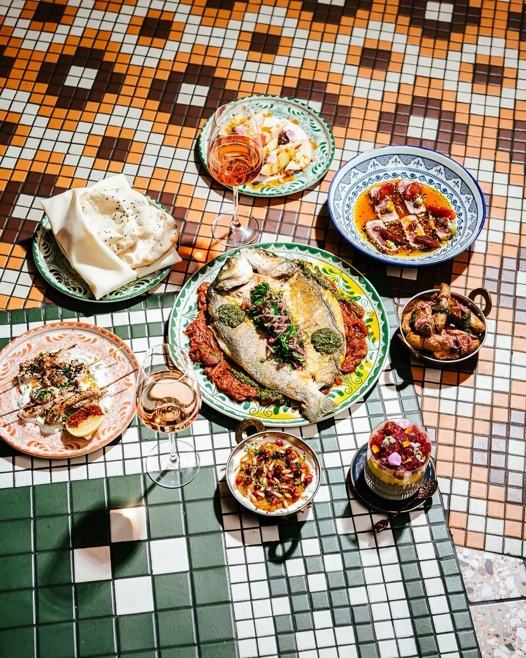 A colorful table setting with various dishes including a whole fish on a large plate, bowls of sauces, a plate of bread, a glass of rosé wine, and small desserts, all on a mosaic tiled table.