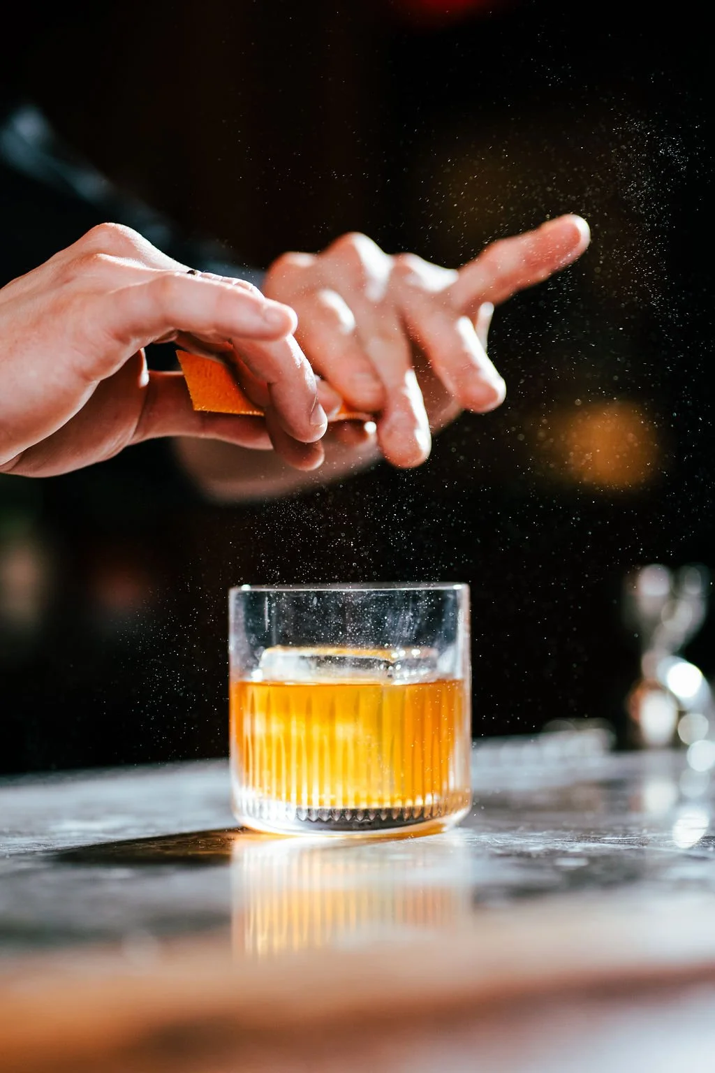 Close-up of a person sprinkling a powder, possibly salt or sugar, into a glass of an amber-colored drink on a wooden surface.