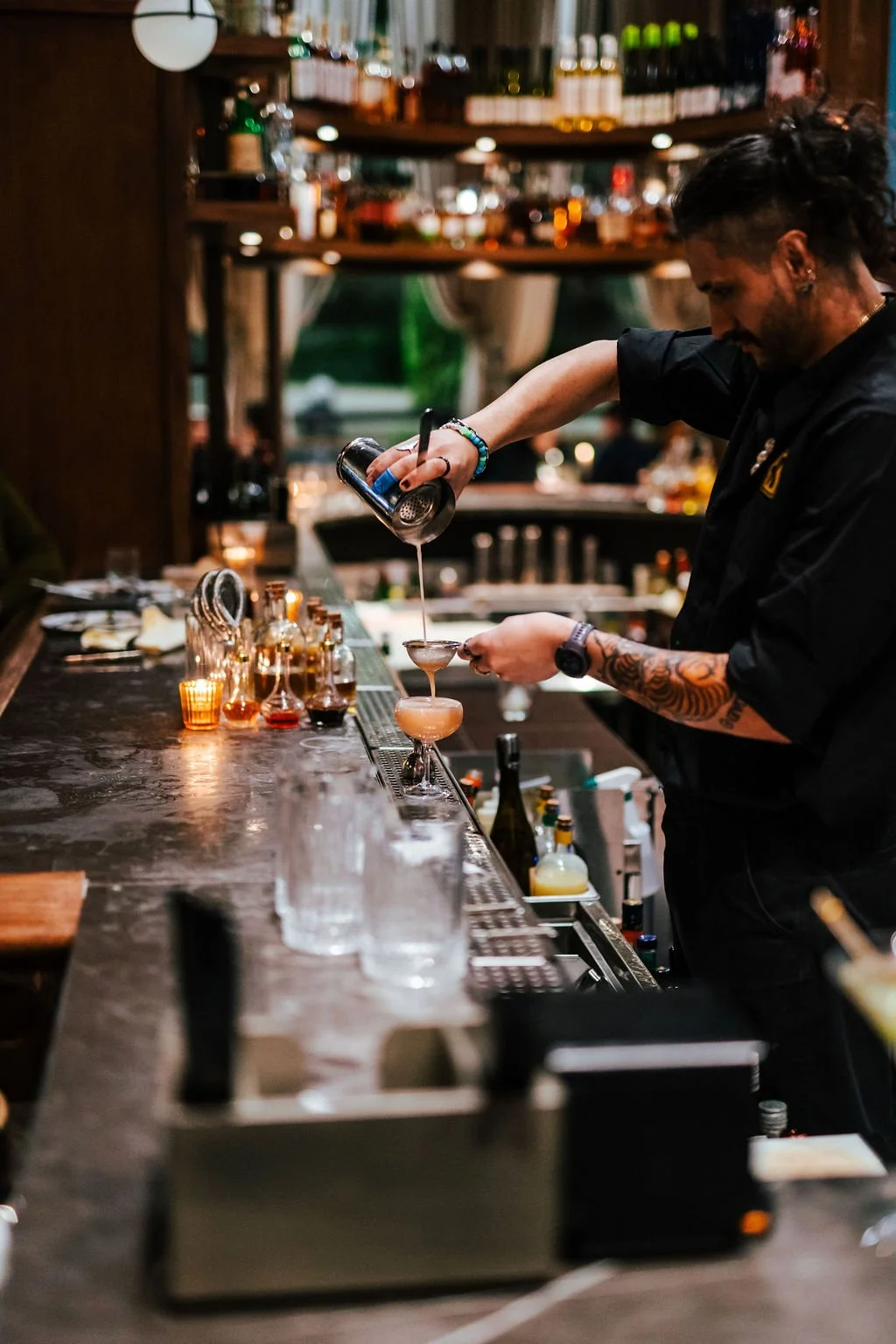 A bartender preparing cocktails behind a bar with various bottles and glasses, pouring a drink into a coupe glass, in a dimly lit bar setting.
