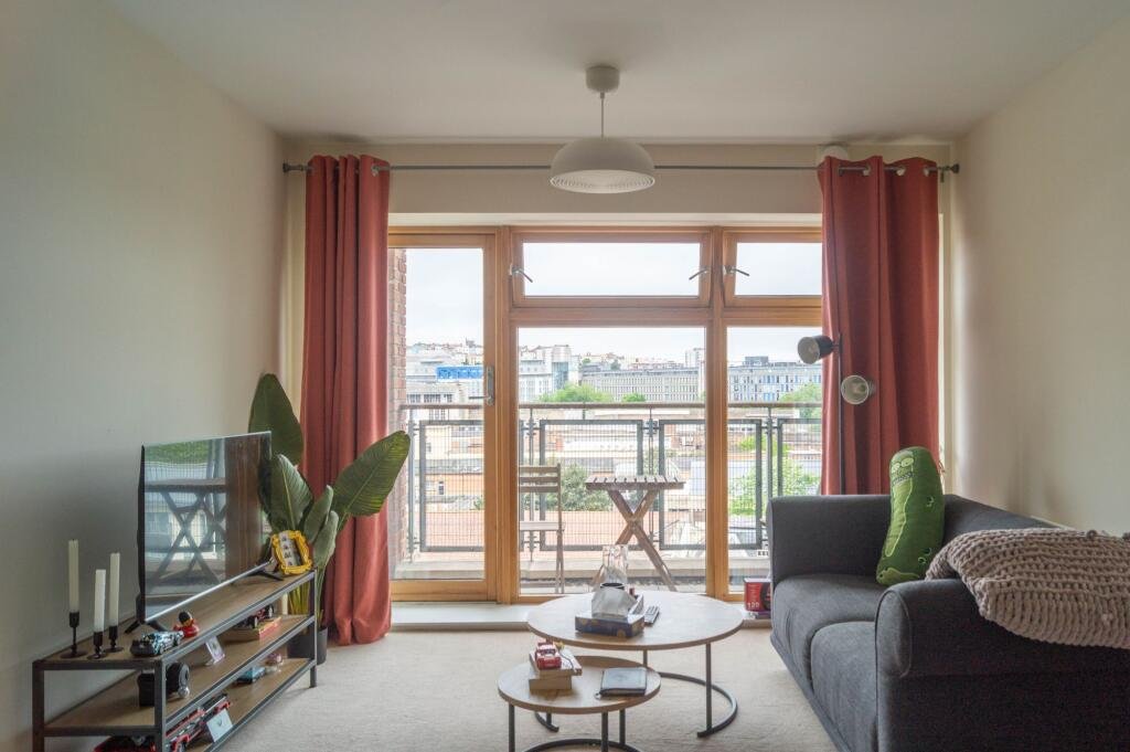 Living room with large window and balcony, gray sofa with a green cushion, coffee table, TV on a stand, and pink curtains.
