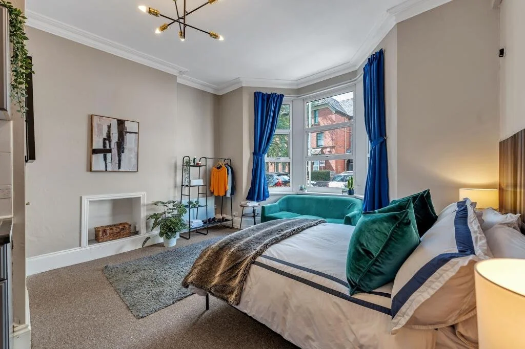 A bedroom with a large bay window, green velvet loveseat, blue curtains, a bed with green pillows, a bedside lamp, and a clothes rack next to a potted plant.