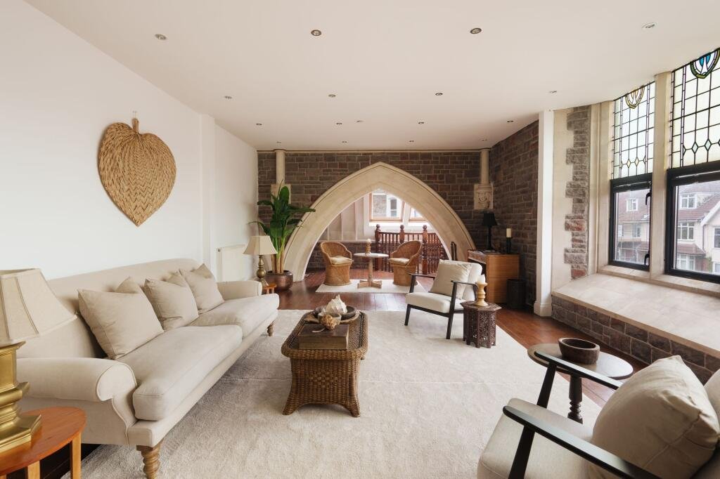 A spacious living room with a white sofa, wicker coffee table, and various chairs, featuring large stained glass windows and a brick accent wall.