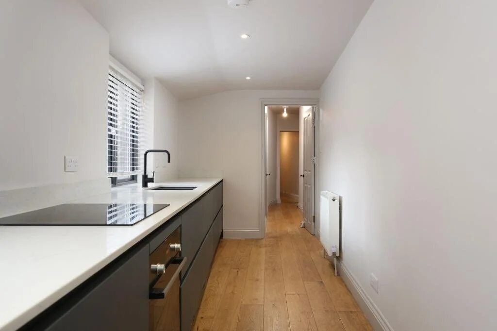 Empty kitchen with white walls, wooden floor, black cabinets, white countertop, stove, sink, window with blinds, and open door leading to a hallway.