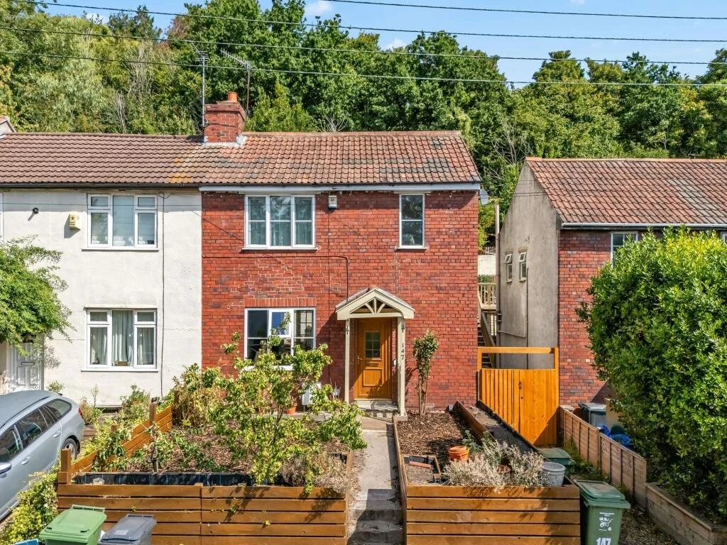 Red-brick two-story house with a wooden front door, small front yard garden, and a neighboring house on each side, surrounded by green trees.