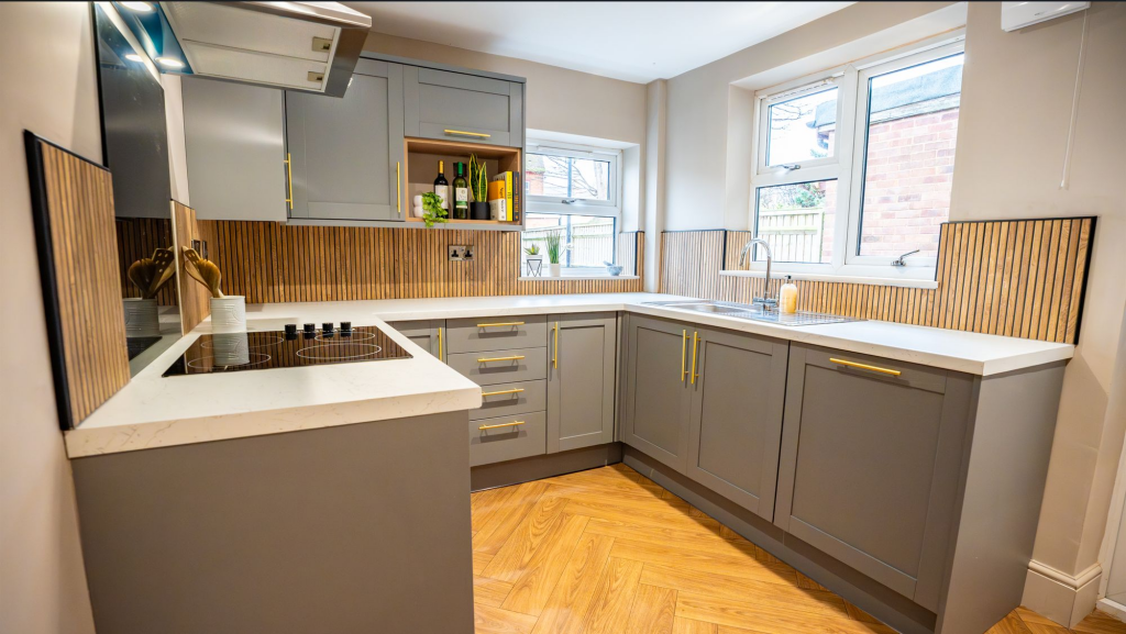 Modern kitchen with gray cabinets, white countertops, wooden backsplash, and hardwood floors. Contains a stove, sink, and shelves with bottles and plants. Natural light from double windows.