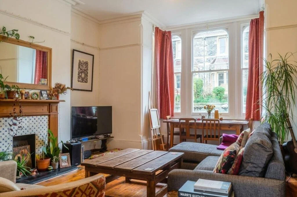 Living room with a large window, corner sofa, wooden coffee table, TV, plants, fireplace, and a dining table with chairs.