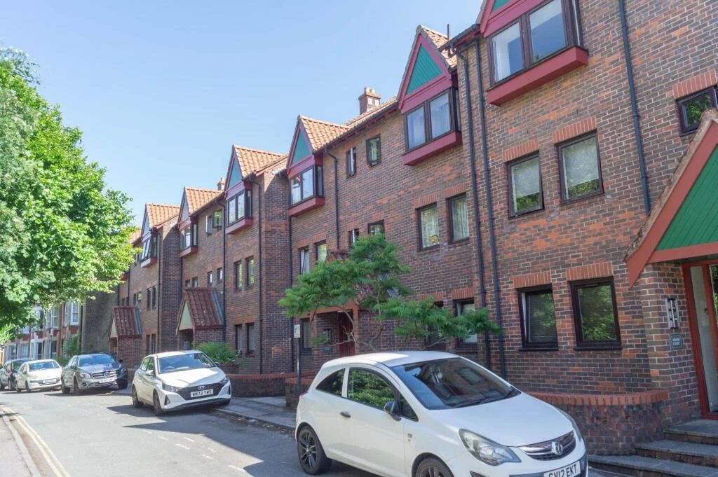 A row of brick multi-story residential buildings with red and green gabled roofs, parked cars on the street, and lush green trees in the neighborhood.