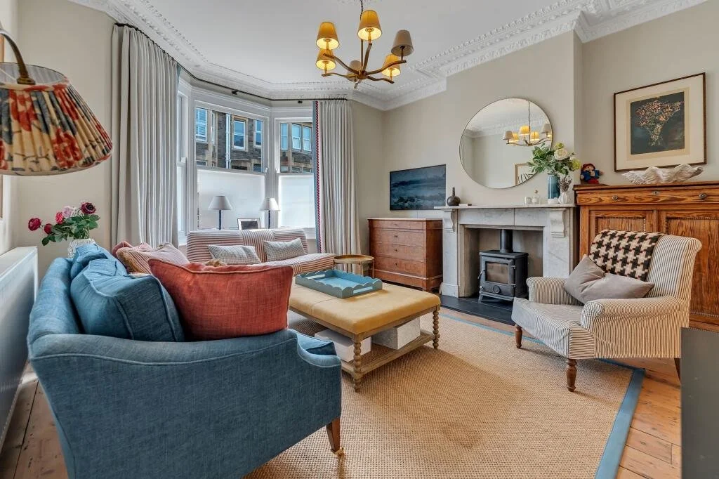 Cozy living room with a blue sofa, pink cushions, beige armchair, and a yellow ottoman in front, a fireplace with a round mirror above, and a wooden sideboard. Large window with curtains, a rug, and a chandelier.