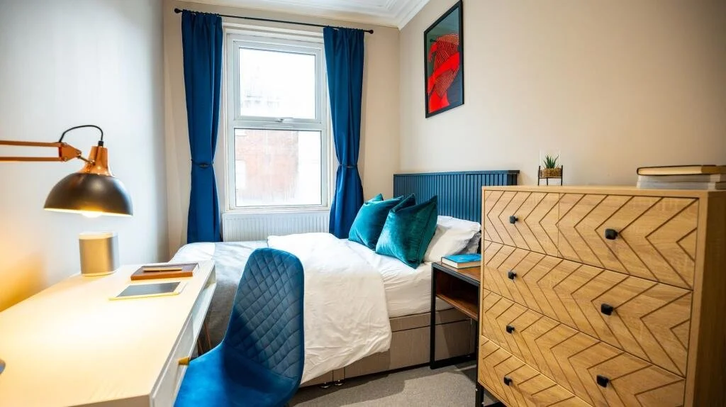 Bedroom with a bed, blue and white decor, a desk with a blue chair, a dresser, and a window with blue curtains.