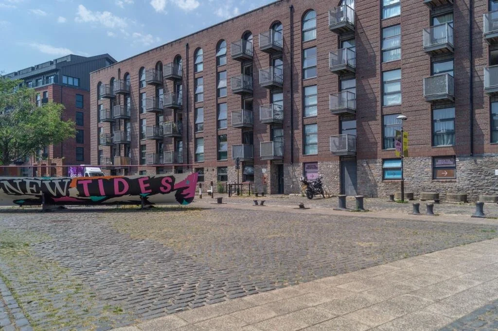 A multi-story brick apartment building with balconies, a sidewalk, a park area with a graffiti sign that says 'NEW TIDES,' a bicycle, several bollards, and a lamppost with a banner on a sunny day.
