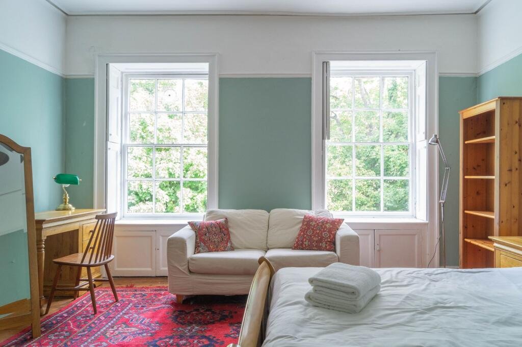 A cozy bedroom with two large windows, a white sofa with two red patterned pillows, a wooden desk with a green lamp, a bed with folded towels, a wooden chair, and a bookshelf. Light green walls with white trim.