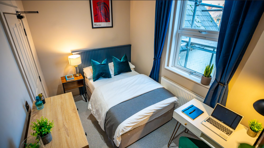 A tidy bedroom with a bed, nightstand, desk with laptop, and window with curtains.