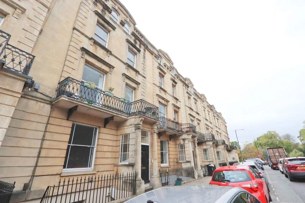 A street view of a three-story historic apartment building with beige stone facade, black wrought iron balconies, and multiple windows. Several cars are parked along the street, and there are trees and streetlights in the background.