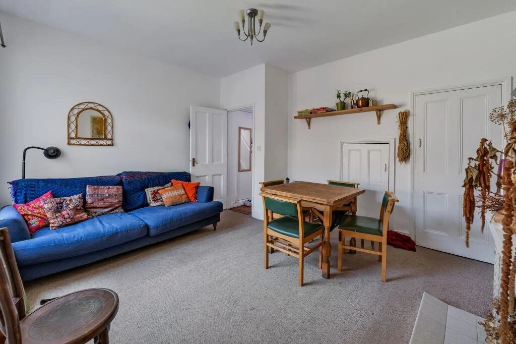 A living room with a blue sofa adorned with multiple colorful pillows, a wooden dining table with four green chairs, a wall-mounted shelf with plants and decor, a closed white door, and a door slightly ajar leading to another room.