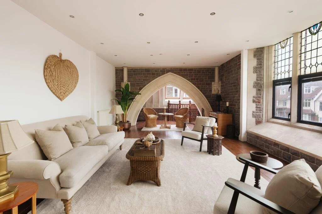 Living room with white sofa, armchairs, wicker coffee table, and large windows with stained glass, exposed brick wall, and a decorative stone archway leading to a small balcony with seating.