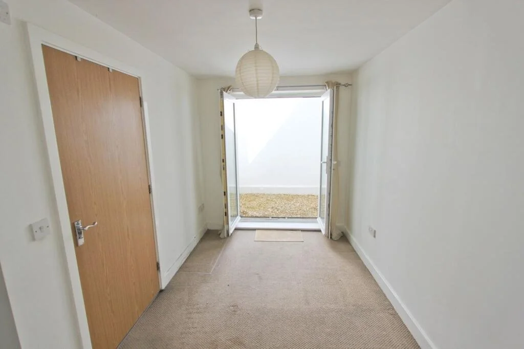 Empty room with beige carpet, white walls, a wooden door, and an open glass door leading outside to a small gravel area.
