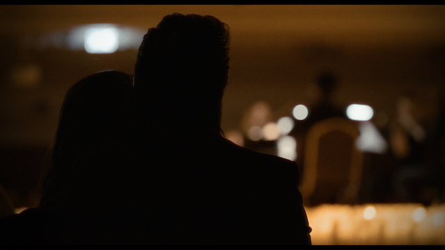 A couple, viewed from behind, watching a performance in a dimly lit venue, focusing on the interaction of the audience with the performers.