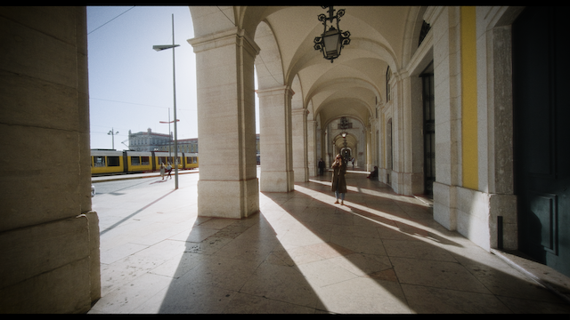 A woman walks alone through a grand arcade, with light casting long shadows on the ground.png