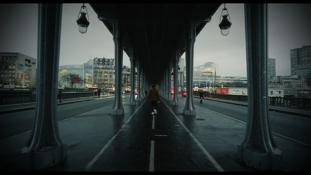 A symmetrical composition of a bridge in a city with a lone cyclist in the middle, the image is under a muted, cloudy sky creating a somber tone with urban architecture in the background.