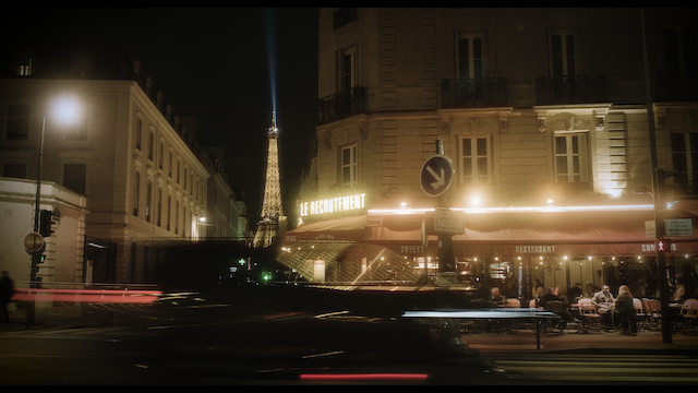 A warmly lit Parisian scene at night featuring the Eiffel Tower in the background, with a foreground of a bustling cafe and streaks of light from passing cars, blending historic charm with lively contemporary life.