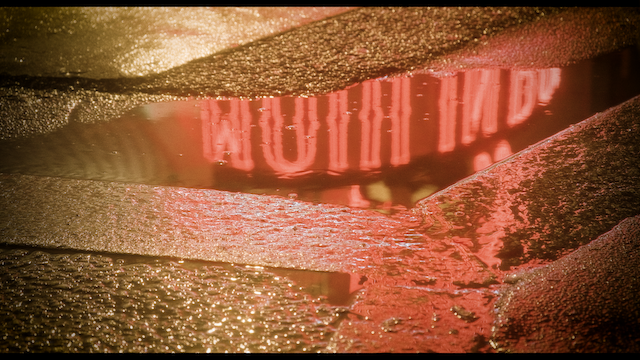 A close-up of a rain puddle reflecting neon lights that spell "MOULIN ROUGE" in distorted, shimmering letters on a wet cobblestone street, creating a moody and atmospheric night scene.