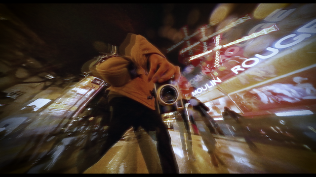 A dynamic, fish-eye lens perspective of a photographer crouching on a wet street at night, capturing the neon-lit Moulin Rouge signage, creating a distorted, wide-angle view of the cityscape.