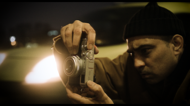 Close-up of a man focusing on adjusting the settings of a vintage camera, highlighting his concentration and the intricate details of the camera against a softly lit background.