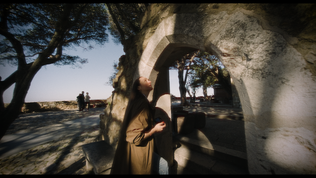 A woman stands in a sunlit stone archway, looking out onto a tree-lined promenade.png