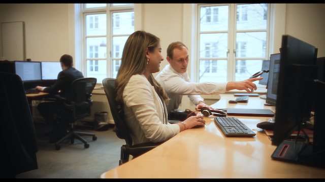 A woman and a man sitting at a desk in an office environment. The man is pointing at the computer screen, explaining something to the woman who is attentively listening.png